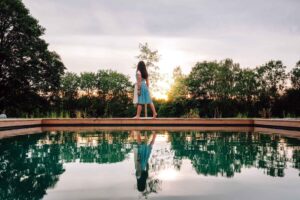 Frau im Dirndl steht am Rand des Biotop-Swimmingpools und betrachtet den Sonnenuntergang im Wellnesshotel Waldeck Bodenmais.